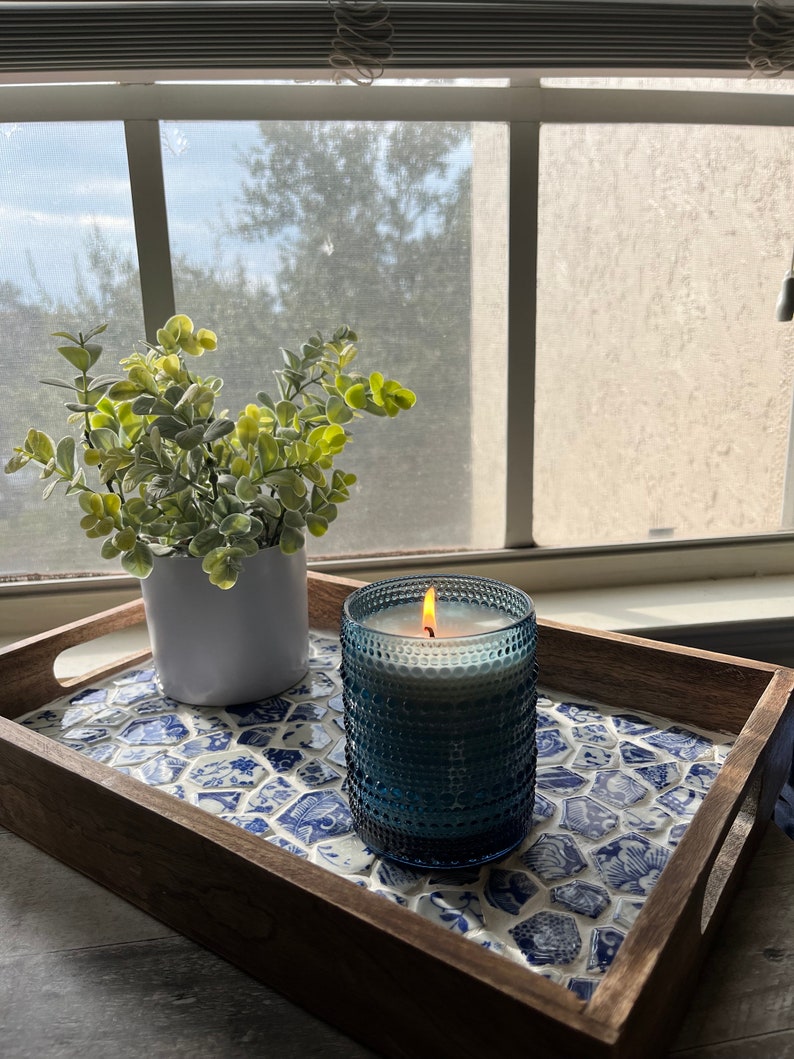 May include: A wooden tray with a blue and white mosaic tile design. A blue glass candle with a white candle inside is lit and sitting on the tray. A small potted plant is also on the tray.