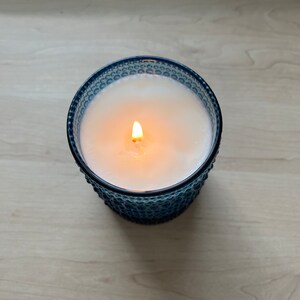 May include: A blue glass candle with a white wax top and a single flame burning in the center.