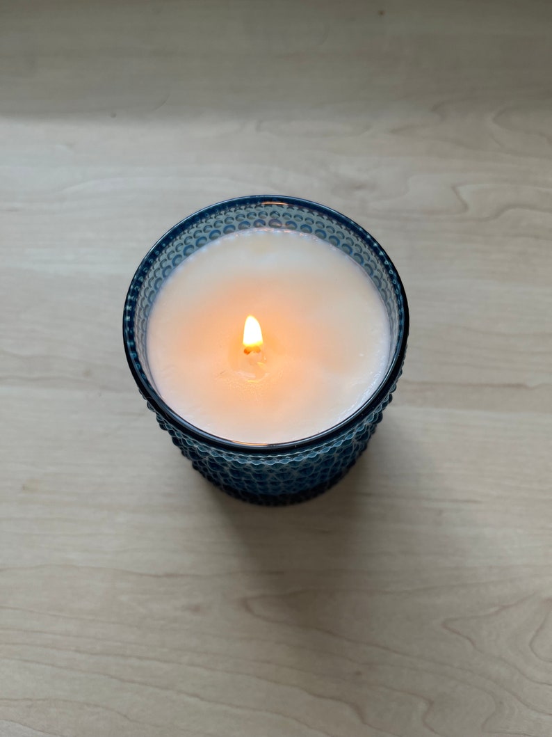 May include: A blue glass candle with a white wax top and a single flame burning in the center.