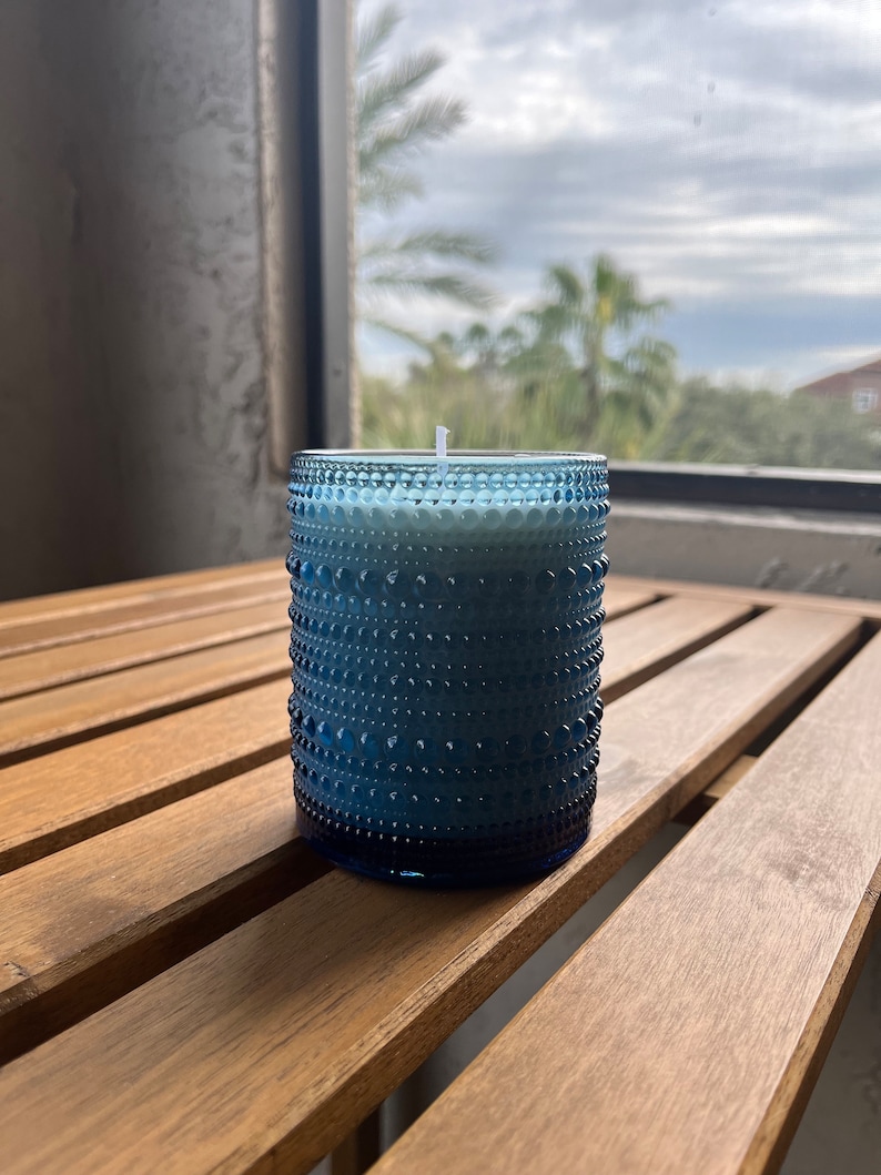 May include: A blue glass candle with a white wax top and a white wick. The candle is sitting on a wooden table.