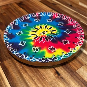 May include: A round wooden serving tray with a tie-dye design. The center features a yellow and white floral pattern, surrounded by a vibrant mix of blue, green, red, and yellow. The edge is trimmed with a white paisley border.