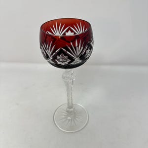 May include: A red and clear cut crystal wine glass featuring a deep ruby red bowl with intricate starburst and geometric patterns cut into the glass, revealing the clear base beneath. The stem is clear glass with a faceted design and a starburst base.