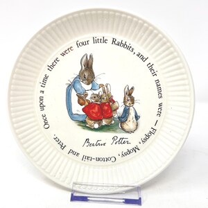 May include: A round, off-white ceramic plate with a ribbed rim featuring a colorful illustration of four rabbits. The text reads: Once upon a time there were four little Rabbits, and their names were - Flopsy, Mopsy, Cotton-tail and Peter. Beatrix Potter.