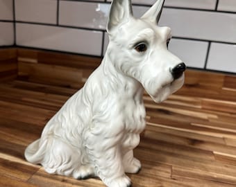 Peut inclure: Figurine en céramique blanche représentant un chien assis, avec un nez noir et des yeux foncés. Le chien a des détails de fourrure et des accents noirs sur les pattes et les oreilles. Objet décoratif.