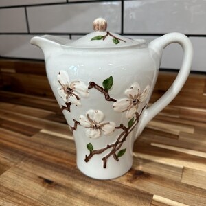 May include: A white ceramic creamer with a lid, handle, and spout. The creamer is decorated with raised white flowers, brown branches, and green leaves. The lid has a small, round knob. The creamer sits on a wooden surface.