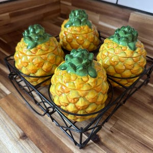 May include: Four ceramic pineapple-shaped containers with green tops and yellow bodies, sitting in a black metal wire basket. The pineapples have a textured surface and are arranged on a wooden surface.
