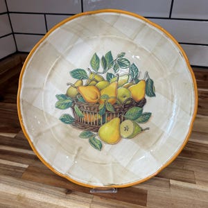 May include: A large, round decorative plate with a cream-colored background and a basket of pears illustration. The plate has a yellow rim and a scalloped edge. The pears are yellow and green, with green leaves.
