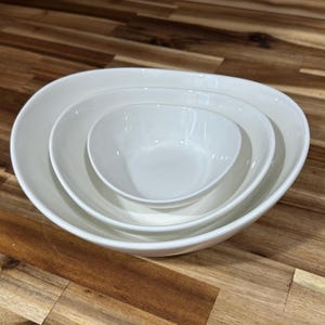 May include: A set of three white ceramic bowls nested together. The bowls are oval-shaped with smooth, glossy surfaces. The largest bowl is at the bottom, followed by a medium-sized bowl, and a small bowl in the center. The bowls are arranged on a wooden surface.