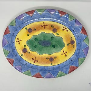 May include: An oval ceramic platter featuring a central green scalloped shape surrounded by a yellow band with small purple floral motifs. The outer border consists of a blue ring with a black swirling line and a perimeter of alternating triangles.