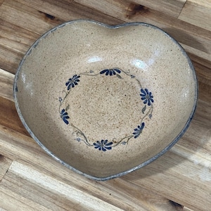 Pode incluir: Tigela de cerâmica em forma de coração com acabamento bege salpicado. O interior apresenta um desenho floral azul em padrão circular. A borda é de um tom bege mais escuro.