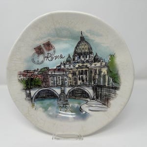 May include: A ceramic plate with a watercolour illustration of Rome, featuring St Peter's Basilica and a bridge. The plate has a light beige background, the word "Roma", and postage stamps. The plate is a decorative souvenir.
