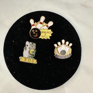 May include: Three metal enamel pins featuring bowling themes. One pin shows bowling pins and a ball with the text BOWLING'S A BALL. Another depicts a green frog holding a bowling pin with the number 500. The third shows pins and a ball with 500 SERIES.