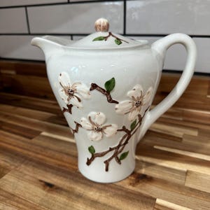 May include: A white ceramic teapot featuring a raised, three-dimensional floral design with brown branches, green leaves, and white blossoms. The teapot includes a matching lid with a small knob, a curved handle, and a spout, all decorated with floral motifs.