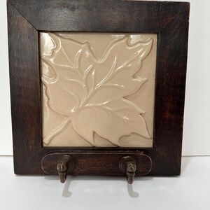 May include: A dark brown wooden key holder with a beige tile featuring a raised leaf design. The key holder has two metal hooks below the tile. The frame is square and the wood has a visible grain.