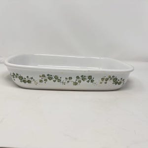 May include: A white rectangular baking dish with rounded corners and a shallow profile. The exterior side features a repeating pattern of green ivy leaves and vines encircling the perimeter of the ceramic vessel, providing a decorative accent.