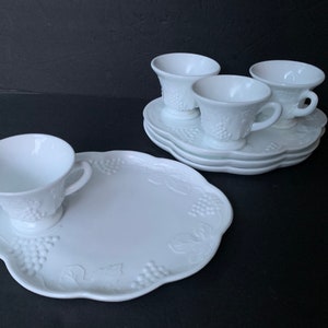 May include: Set of four white milk glass cups and saucers with a grape design. The cups have a footed base and the saucers have a scalloped edge. There is also a larger white milk glass plate with a grape design and a scalloped edge.