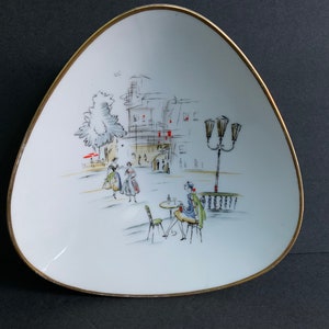 以下が含まれることがあります： 金色の縁取りのある白い三角形のセラミック皿。皿には、歩いている人やカフェに座っている人など、街の風景が白黒で描かれています。