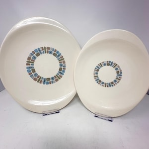 May include: Two off-white ceramic plates of different sizes, each featuring a central circular pattern composed of small, repeating rectangular shapes in shades of blue, brown, and black. The plates have a smooth, glossy finish and a wide rim.