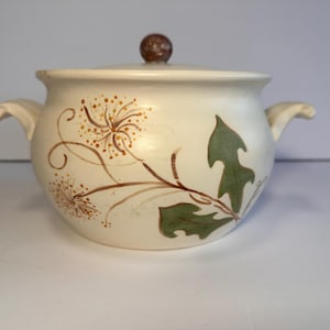 May include: A white ceramic casserole dish with a lid. The dish has a floral design with brown and green leaves and yellow flowers.