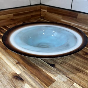 May include: A decorative glass bowl with a wide, shallow design. The bowl features a gradient of colors, with a dark brown rim, a white band, and a light blue center. The bowl is likely used for decorative purposes.