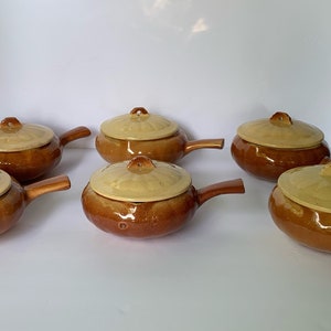 May include: Six brown ceramic casserole dishes with lids. The lids have a light yellow glaze and a raised design. The dishes have a brown glaze and a handle on one side.