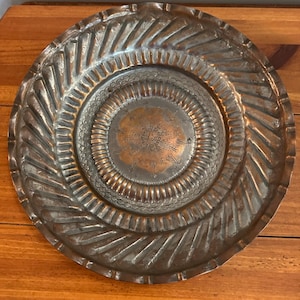 May include: A round copper tray featuring a central medallion with etched floral patterns, surrounded by a ring of scalloped detailing. The outer rim displays a series of diagonal, embossed ridges that extend toward the edge of the metal plate.