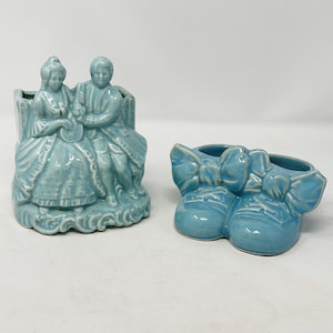 May include: A light blue ceramic planter featuring a sculpted couple sitting on a bench and a separate light blue ceramic planter shaped like a pair of baby shoes with large bows on the front. Both items have a glossy finish and visible crazing.