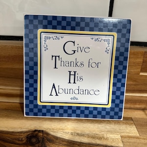 May include: Square decorative tile with the text "Give Thanks for His Abundance." The tile has a white center with the text in dark blue, framed by a yellow border. The outer border is a blue and white checkered pattern.