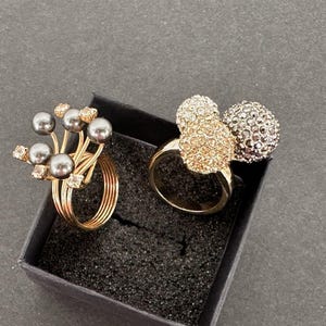 May include: Two rings displayed in a black box. One ring features a cluster of gray pearls and clear crystals on a gold band. The other ring has a gold band with three crystal-covered spheres, two in gold and one in silver.