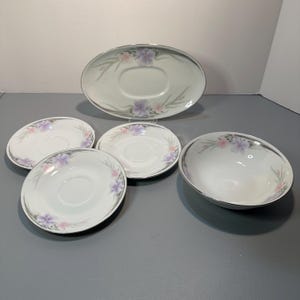 May include: A white porcelain dish set with a floral design. The set includes four saucers, one oval serving dish, and a small bowl. Each piece has a silver trim and a pattern of purple and pink flowers.