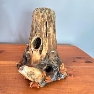 May include: A ceramic sculpture shaped like a tree stump with a textured brown and tan bark finish. The piece features two hollow openings and is decorated with small, orange-colored sculpted fungi or leaves attached to the base and sides.