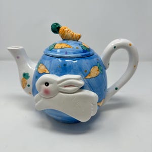 May include: A ceramic teapot with a blue background and a white bunny design. The teapot has a carrot on top of the lid and carrot and leaf designs on the body. The handle and spout are white with orange and yellow polka dots and carrot designs.
