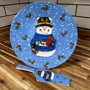 May include: A blue ceramic snowman plate and matching server. The plate features a snowman wearing a blue scarf and holding a gift. The plate and server are decorated with holly and snowflakes. The server has a matching snowman design.