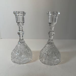 May include: Two clear glass candle holders. Each has a detailed, floral-patterned base and a slender stem leading to a cup-shaped top. The candle holders are made of clear glass and are designed to hold candles.