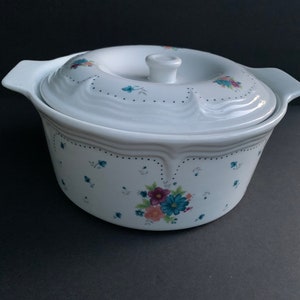 Könnte beinhalten: Weiße Keramik-Auflaufform mit Deckel. Die Form hat ein Blumenmuster mit blauen, rosa und violetten Blumen. Der Deckel hat einen Knopf oben.