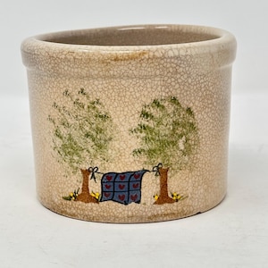 May include: A cylindrical ceramic crock with a beige crackle glaze finish. The exterior features a hand-painted scene of a blue quilt with red hearts hanging on a clothesline between two green trees with brown trunks and small yellow flowers.