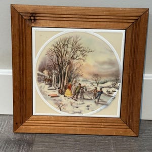 May include: A framed tile featuring a winter scene with people ice skating and playing hockey. The artwork is set within a light-colored circular design, surrounded by a wooden frame. The scene includes trees, snow, and a building.