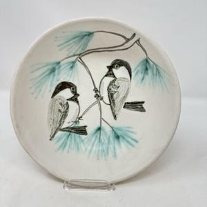 May include: A round white ceramic bowl featuring a hand-painted illustration of two small birds perched on a pine branch. The birds are rendered in black and grey, while the pine needles are painted in a soft teal green color.