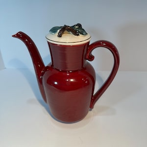 May include: A glossy, red ceramic teapot with a long spout and a curved handle. The lid is off-white with a decorative green and brown floral design. The teapot is a classic design.