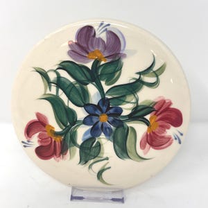 May include: A round ceramic plate with an off-white base featuring a hand-painted floral design. The center displays a small blue flower with a yellow center, surrounded by larger purple and pink flowers with green leaves and stems.