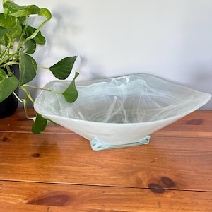 May include: A square-shaped glass bowl featuring a translucent pale green and white marbled swirl pattern. The bowl has a wide, open top with gently undulating edges and rests on a small, clear glass base, showcasing a smooth, polished finish.