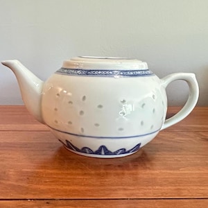 May include: A white ceramic teapot featuring a blue geometric pattern around the rim, a blue leaf-like border at the base, and a subtle embossed dot texture across the body. The teapot includes a matching lid, a curved spout, and a handle.