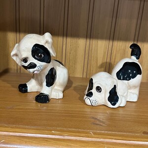 May include: Two ceramic puppy figurines. One is sitting with its head tilted, and the other is lying down. Both are white with black spots and accents. The figurines are on a wooden shelf.