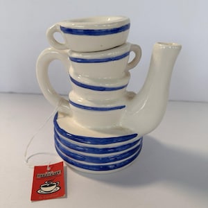 May include: A white ceramic teapot with blue stripes, featuring a stacked design with a cup on top. The teapot has a curved handle and spout. A red tag with the text "Chez Cafe" is attached.