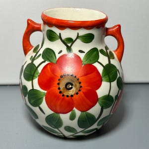 May include: A small, ceramic vase with a white base, adorned with a large red flower and green leaves. The vase has two orange handles and a matching orange rim. The floral design is hand-painted.