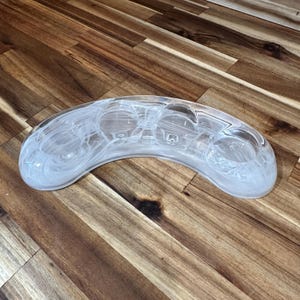 May include: A clear, curved glass candle holder with a white, swirled design. The holder has three circular spaces for candles and is set on a wooden surface. The overall shape is a rounded crescent.