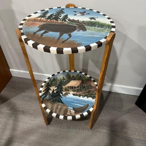 May include: A two-tiered wooden side table with hand-painted nature scenes. The top tier features a moose in a lake, while the bottom tier depicts a cabin by a lake. The edges are decorated with a brown and white checkerboard pattern.