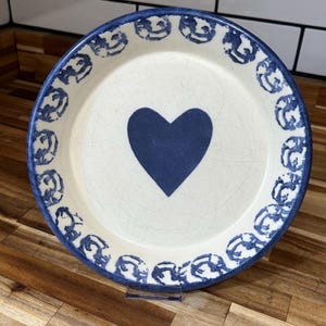 May include: A round, off-white ceramic plate with a navy blue heart in the center. The rim features a repeating pattern of blue decorative elements. The plate has a slightly distressed appearance, adding to its charm.