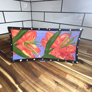 May include: A rectangular glass tray featuring a hand-painted design of two large orange lilies with green leaves against a light blue background. The tray has a black border decorated with white polka dots along the entire outer edge.