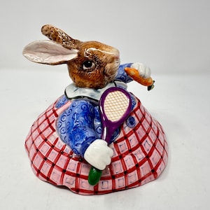 May include: A ceramic figurine of a brown rabbit wearing a blue patterned jacket and a pink and red plaid skirt. The rabbit holds a purple tennis racket in one hand and an orange carrot in the other, featuring white gloves and a white collar.